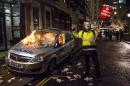 An anti-capitalist protester wearing a Guy Fawkes mask holds a placard as he stands alongside a burning police car during the "Million Masks March", organised by the group Anonymous, in London on November 5, 2015