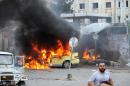 In this photo released by the Syrian official news agency SANA, Syrians gather in front of a burning car at the scene where suicide bombers blew themselves up, in the coastal town of Tartus, Syria, Monday, May 23, 2016. Syrian TV said suicide bombers blew themselves followed by a car bomb in a parking lot packed during morning rush hour. (SANA via AP)